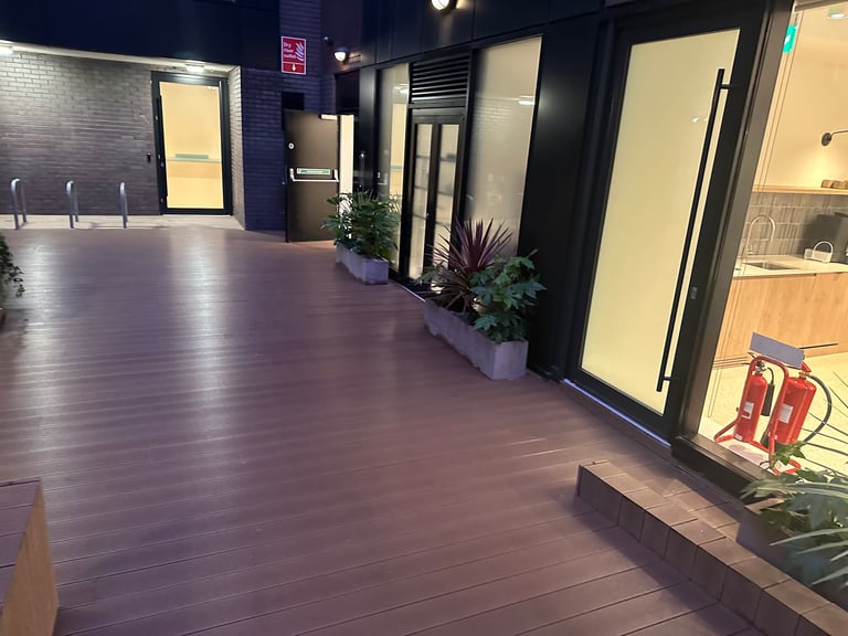 Empty modern hallway with wooden flooring, dark walls, and illuminated frosted glass doors leading to storefronts and potted plants