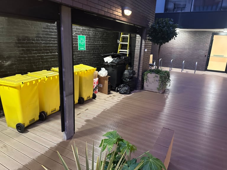 Outdoor patio area with yellow waste bins, wooden deck, green plant, black brick wall, and yellow ladder at night