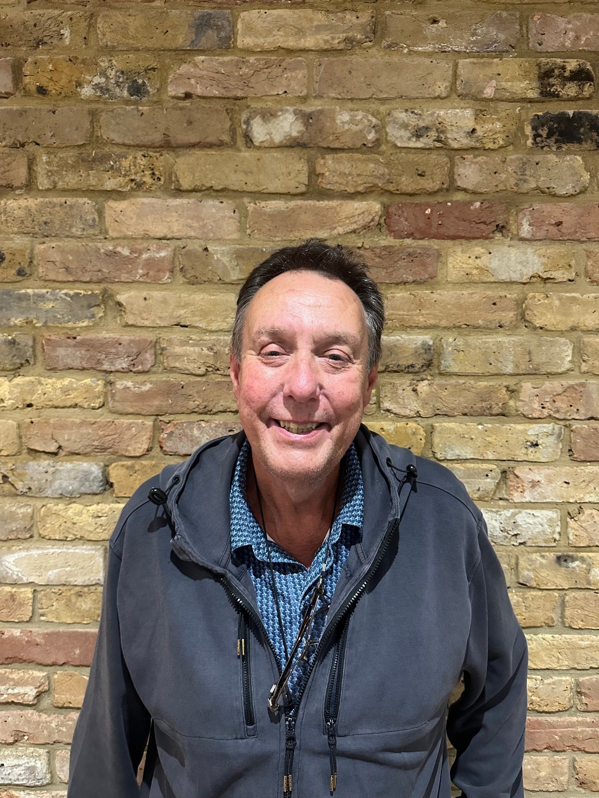 Smiling man wearing a dark jacket over a blue checkered shirt, posing in front of a weathered brick wall.