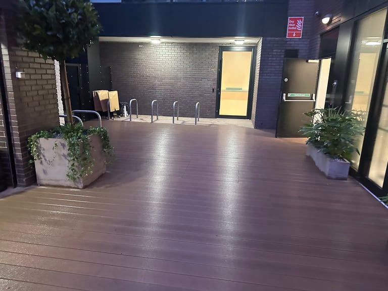Empty modern outdoor deck with purple-toned flooring, planters with greenery, and illuminated building entrance with doors