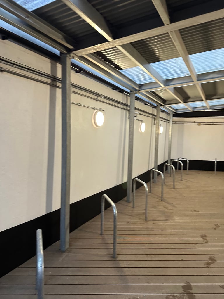 Empty modern shelter or covered walkway with metal frame, white walls, glass roof panels, and a black bench along the side