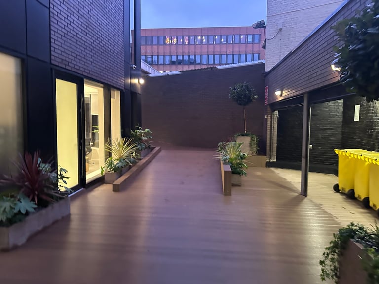 Evening view of a modern urban courtyard with illuminated building entrance, planters, and yellow barriers along wet pavement